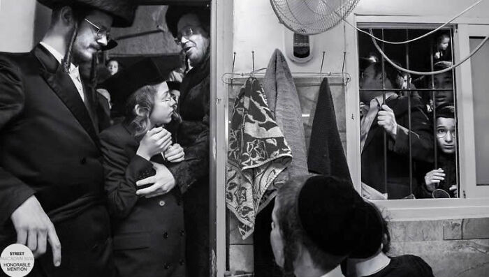 Black and white street photo showing a group of people interacting in a confined space with bars on the window.