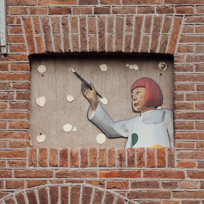 Street art interpretation of public spaces showing a woman with a red bob haircut painting on a building wall.
