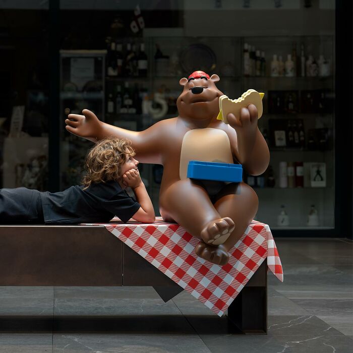 Child lying next to a large bear sculpture eating a sandwich, showcasing clever street art interpretations of public spaces.