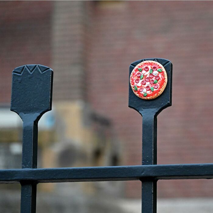 Street art on a black metal fence post featuring a small, detailed pizza decoration in a public space.