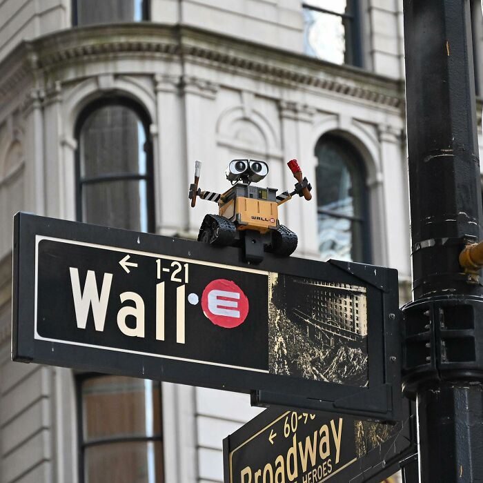 Street art interpretation featuring a Wall-E figure creatively placed on a Wall Street sign in public space.