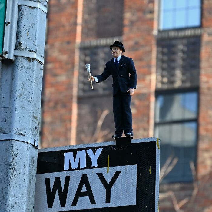 Miniature street art figure holding a paintbrush on a traffic sign, showcasing clever interpretations of public spaces through street art.