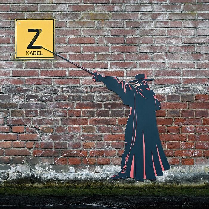 Street art on a brick wall showing a figure in a cloak holding a cable connected to a yellow sign, creative public space artwork.