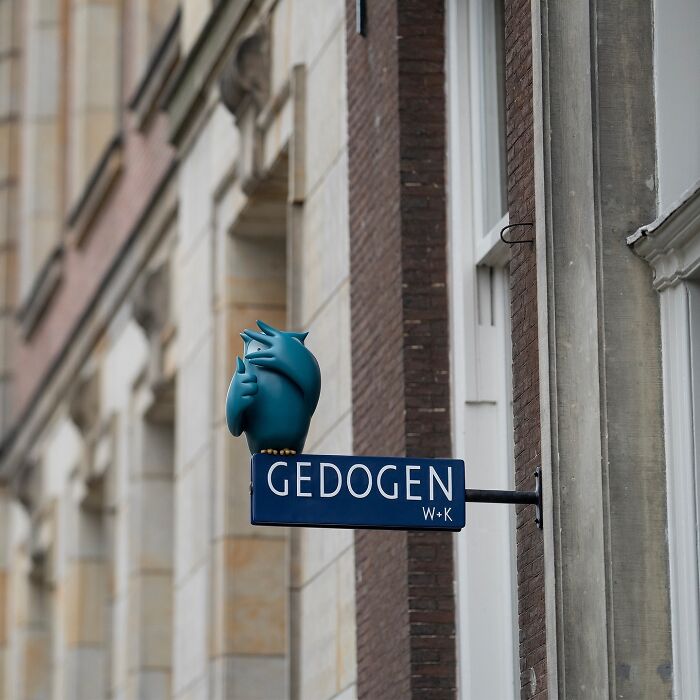 Street art interpretation of public spaces featuring a blue bird sculpture on a building sign in an urban setting.