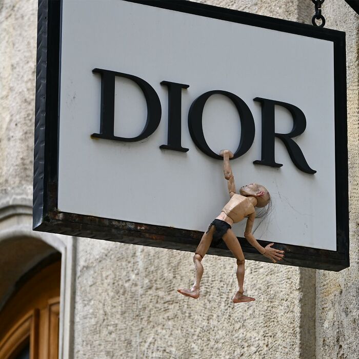 Street art interpretation of public spaces showing a small puppet climbing a Dior sign on a city wall.
