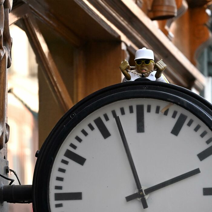 Small street art figure with sunglasses and a cap placed on top of a public clock in an urban setting.