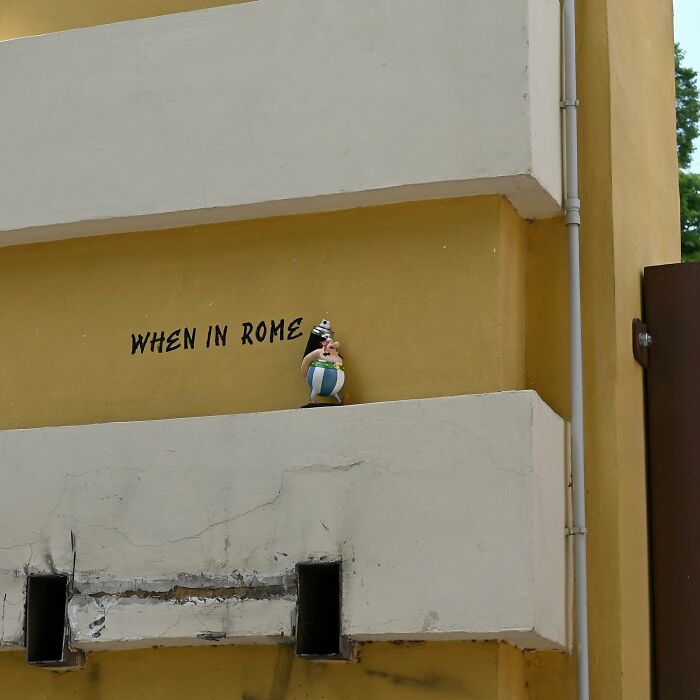 Street art featuring a small figurine on a ledge and the phrase when in Rome on a yellow public space wall.