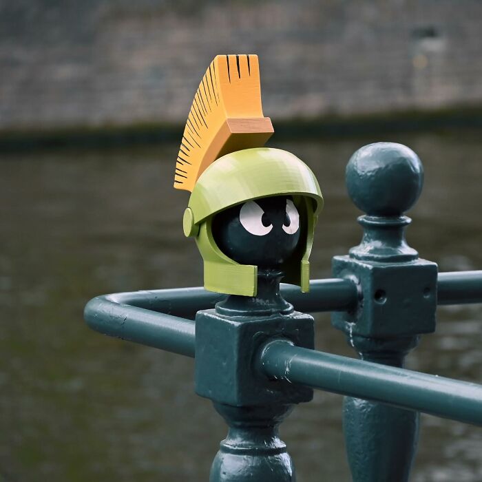 Street art interpretation of public spaces showing a small figure with a yellow helmet on green metal railing by water.