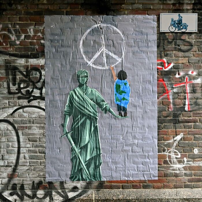 Street art on a brick wall depicting Lady Justice holding a child painting a peace symbol, showcasing clever public space interpretations.
