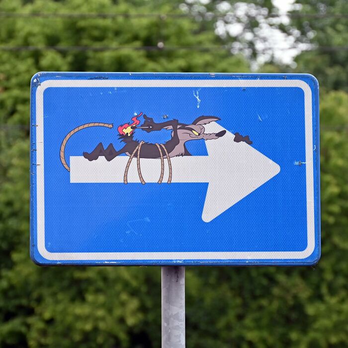 Street art creatively transforming a public space sign with a cartoon character interacting with an arrow on a blue background.