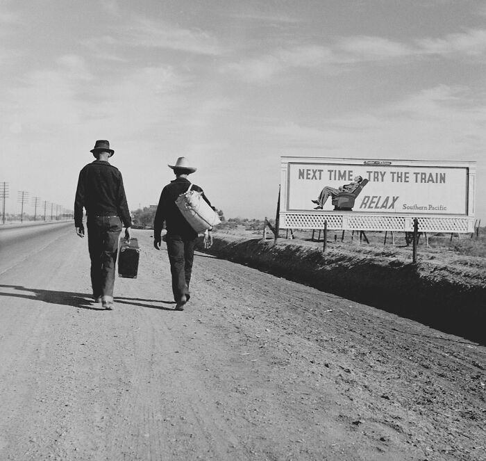 Two men walking on a deserted dirt road past a billboard in a somber photo from history telling a gut-wrenching story.