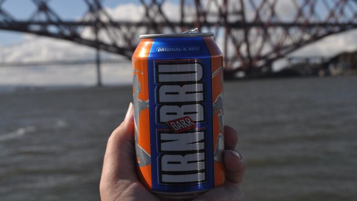 Hand holding an IRN BRU can by a river with a bridge in the background, illustrating ingenious inventions repurposed.