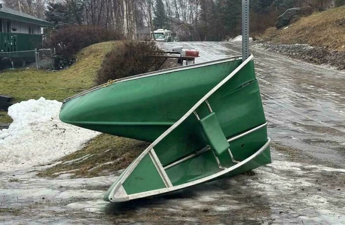 Green boat lifted and twisted on icy road showcasing wild Mother Nature doing things on its own terms in a residential area.