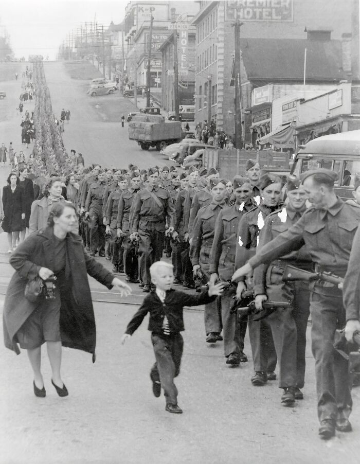 Line of soldiers marching in a city street with a woman and child reaching out, a somber moment captured in history.