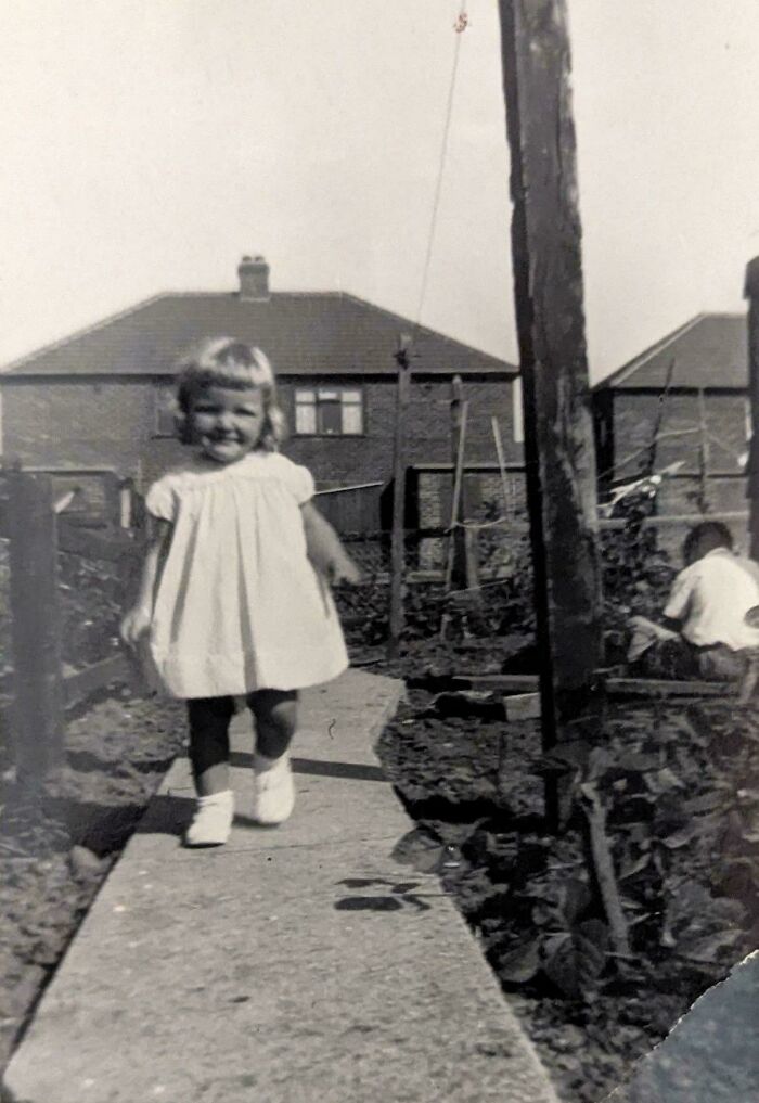 Black and white candid glimpse into the past showing a smiling young girl walking on a garden path near houses.