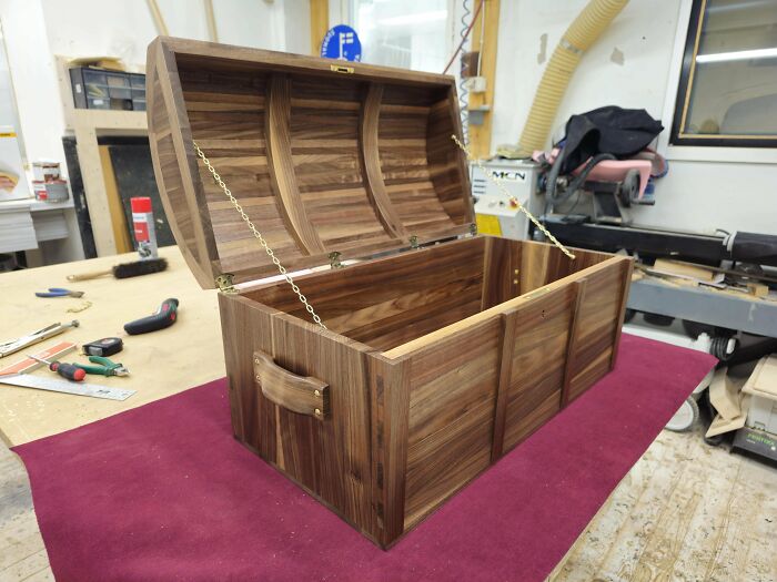 Handcrafted wooden chest with curved lid and brass chains showcasing fine woodworking skills in a workshop setting.
