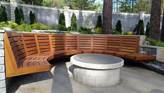 Curved wooden bench crafted by woodworkers surrounding a stone fire pit in an outdoor garden setting.