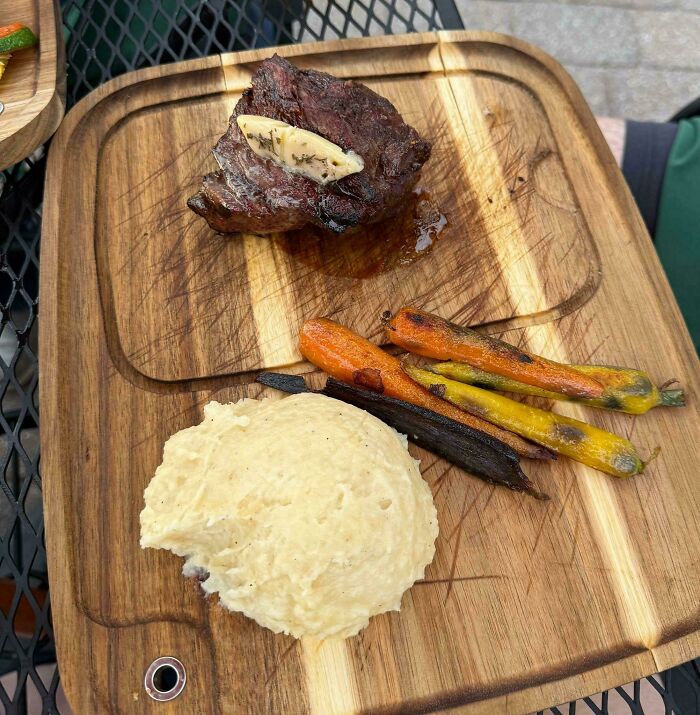 Steak with butter, charred baby carrots, and mashed potatoes served on a wooden cutting board in a restaurant.