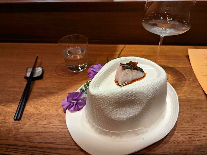 A piece of fish served on a white textured hat-shaped plate with purple flowers at a restaurant.