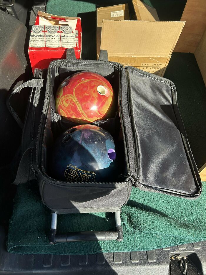 Black and red bowling balls in a carrying case among cardboard boxes dumpster diving treasures found outdoors
