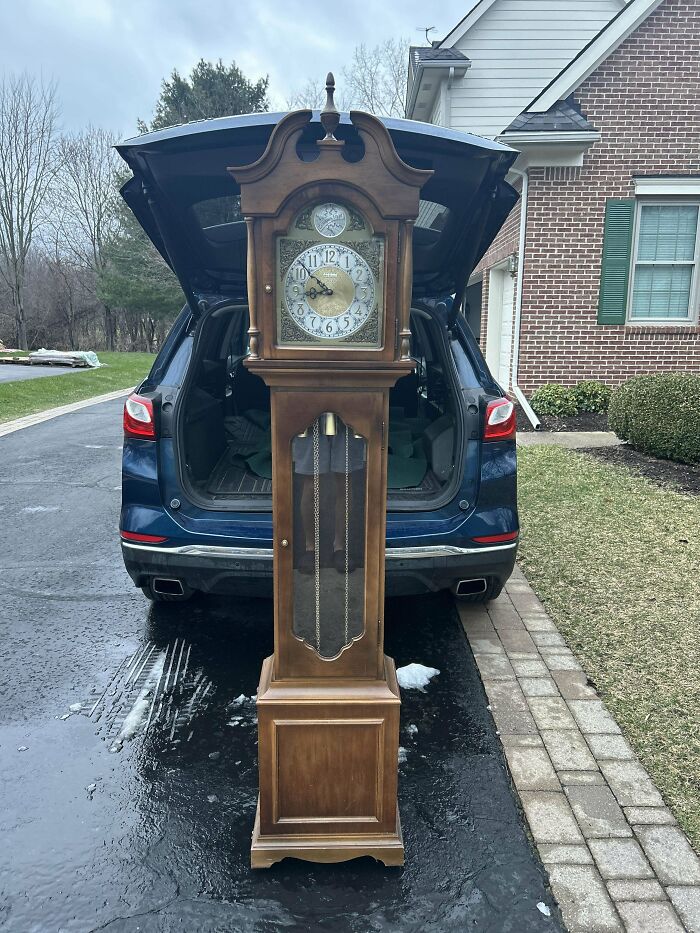 Antique grandfather clock treasure found by dumpster diving standing in front of open car trunk on driveway.