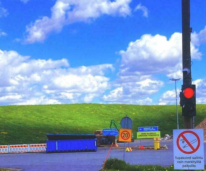 Traffic light showing red at road with signs and a green hill under a partly cloudy sky in an interesting scene.