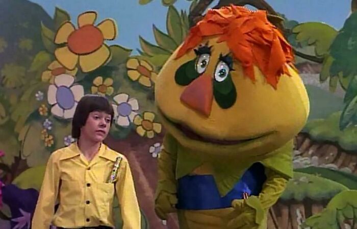 Child and large puppet character in colorful nostalgic TV show set with oversized flowers, evoking old age memories.