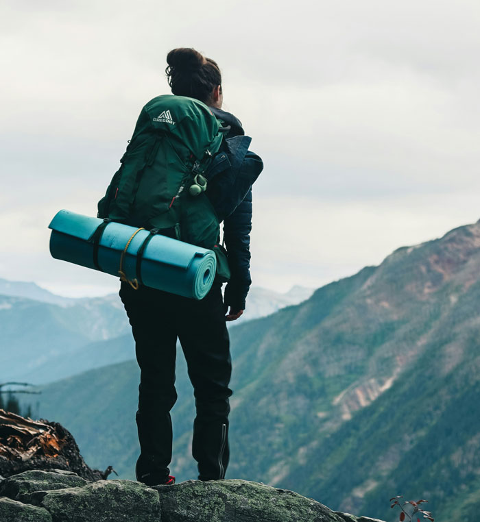 Hiker with backpack and sleeping pad overlooking mountains, illustrating tips to stay safe and alive outdoors.