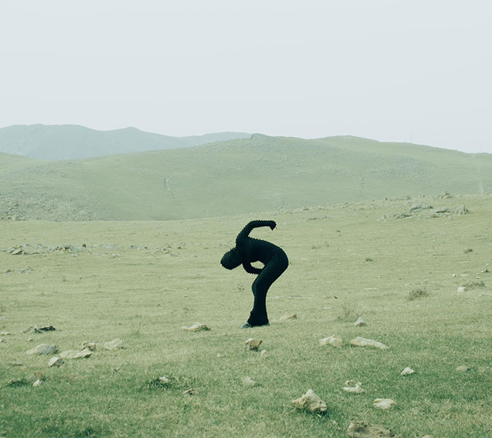 Person in full-body cat suit crawling on grassy field with hills, embodying mysterious figure terrorizing neighborhood at night.