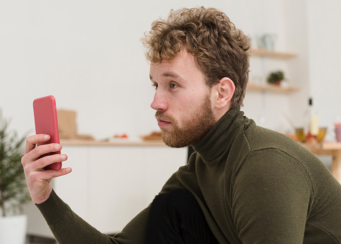 Man in a green turtleneck sitting indoors, intently looking at his red smartphone, revealing employee secrets. - 7