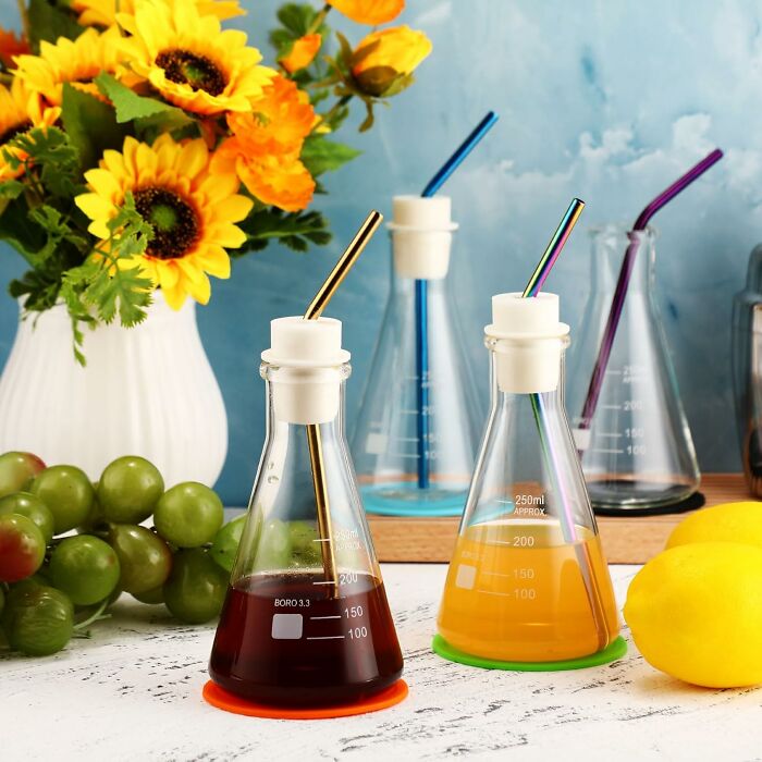 Colorful glass beakers with metal straws holding drinks, surrounded by fruit and sunflowers, showcasing peak coolness items.
