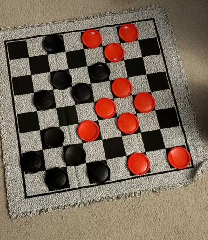 Checkerboard game with red and black pieces on a woven Prime Day treasures mat on carpeted floor.
