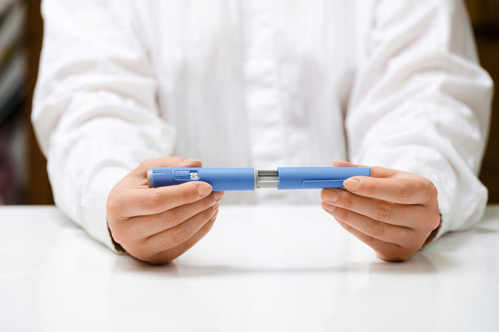 Person in white shirt holding blue weight loss medication injector pen with both hands over a white table. Person in white shirt holding blue weight loss medication injector pen with both hands over a white table.