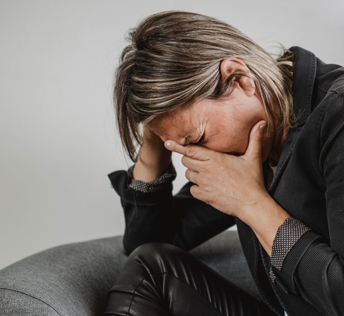 Woman in black jacket sitting on a gray couch, holding her head in distress, reflecting the unfair struggle of a girl surviving cancer.