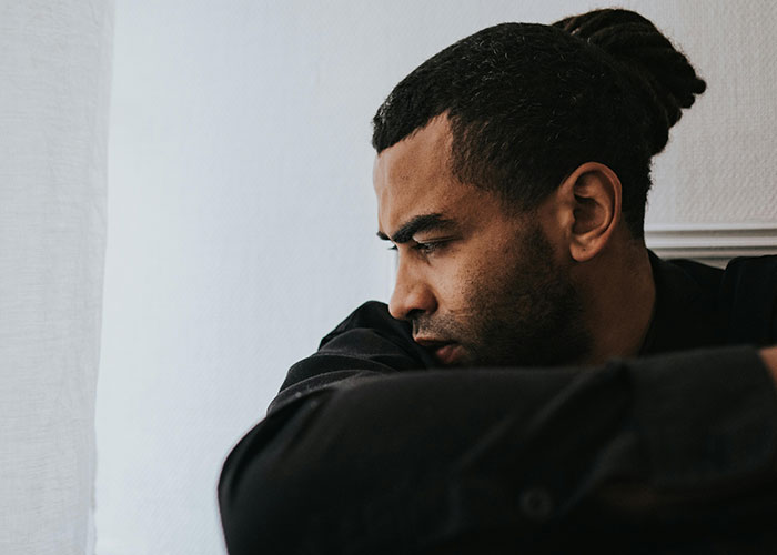 Man with worried expression and dreadlocks, sitting alone near a window, reflecting on worst cheating stories.