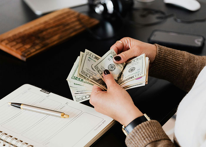 Hands counting US dollar bills at desk with notebook and pen, illustrating micromanagement in the workplace.