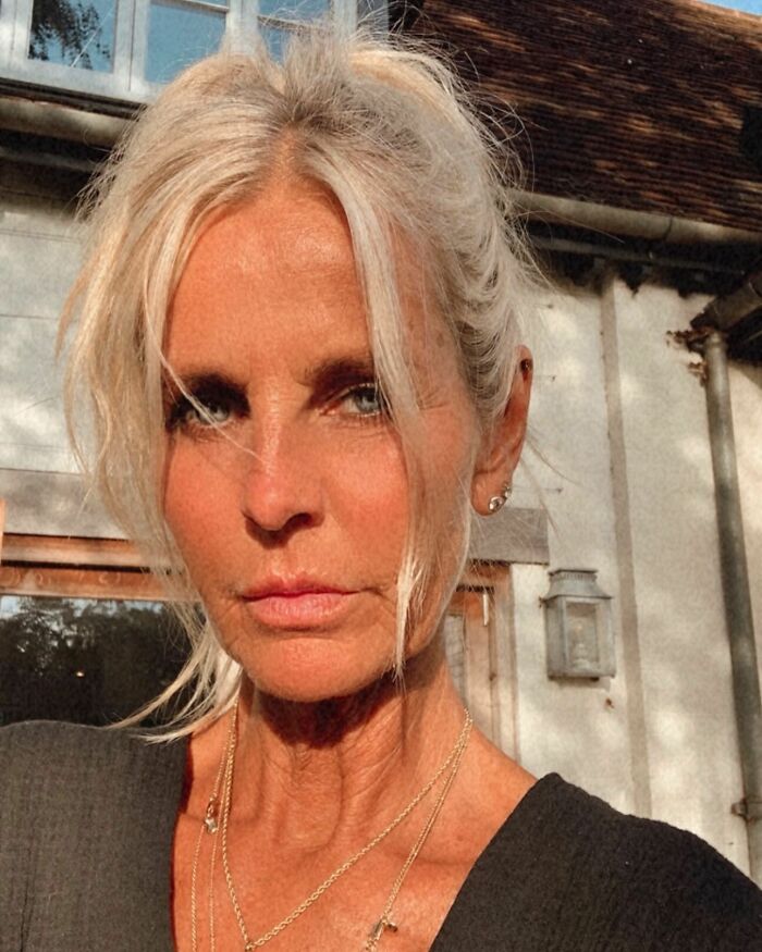 Ulrika Jonsson outdoors with a tanned face, light hair pulled back, wearing delicate gold necklaces and a dark top.