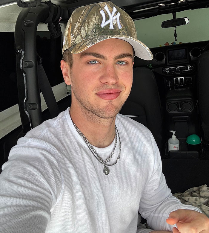 Young man wearing a camouflage cap and white shirt taking a selfie inside a car, related to Sydney Sweeney American Eagle jeans. - 1