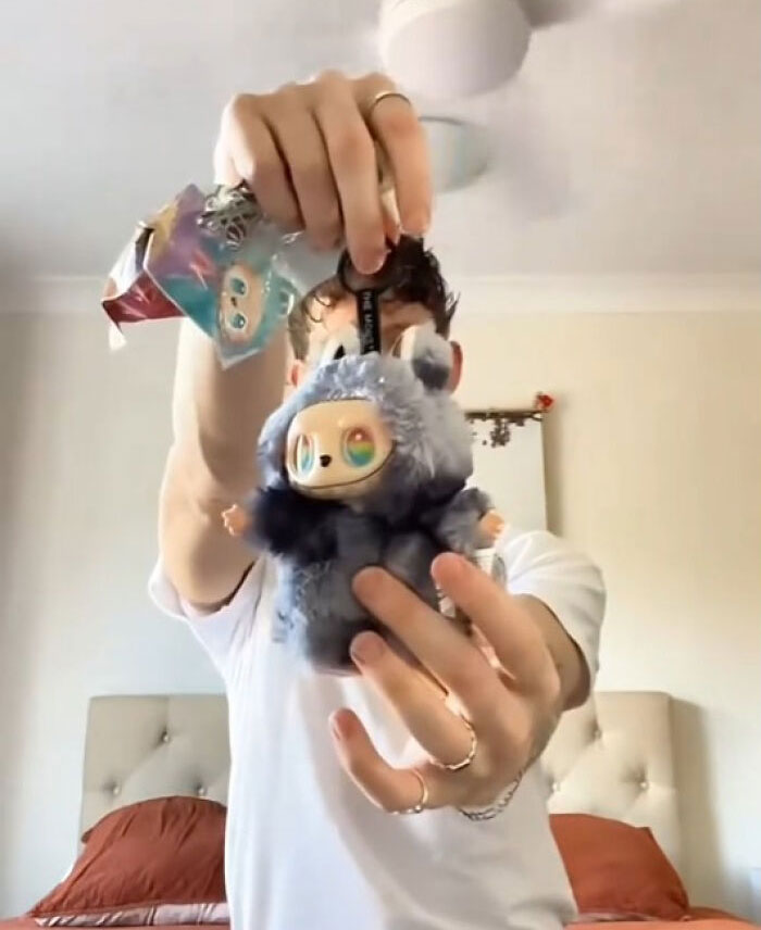 Person holding creepy Labubus doll fakes with large colorful eyes in a bedroom with a ceiling fan and bed in background.