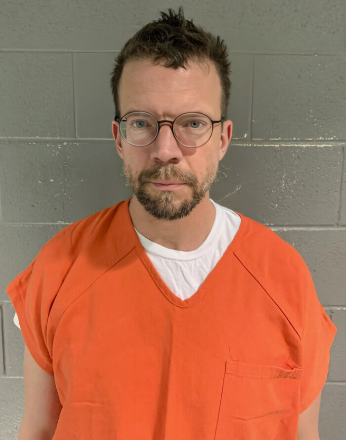 Man in orange prison jumpsuit and glasses standing against a gray wall illustrating consequences of job loss from stupidity