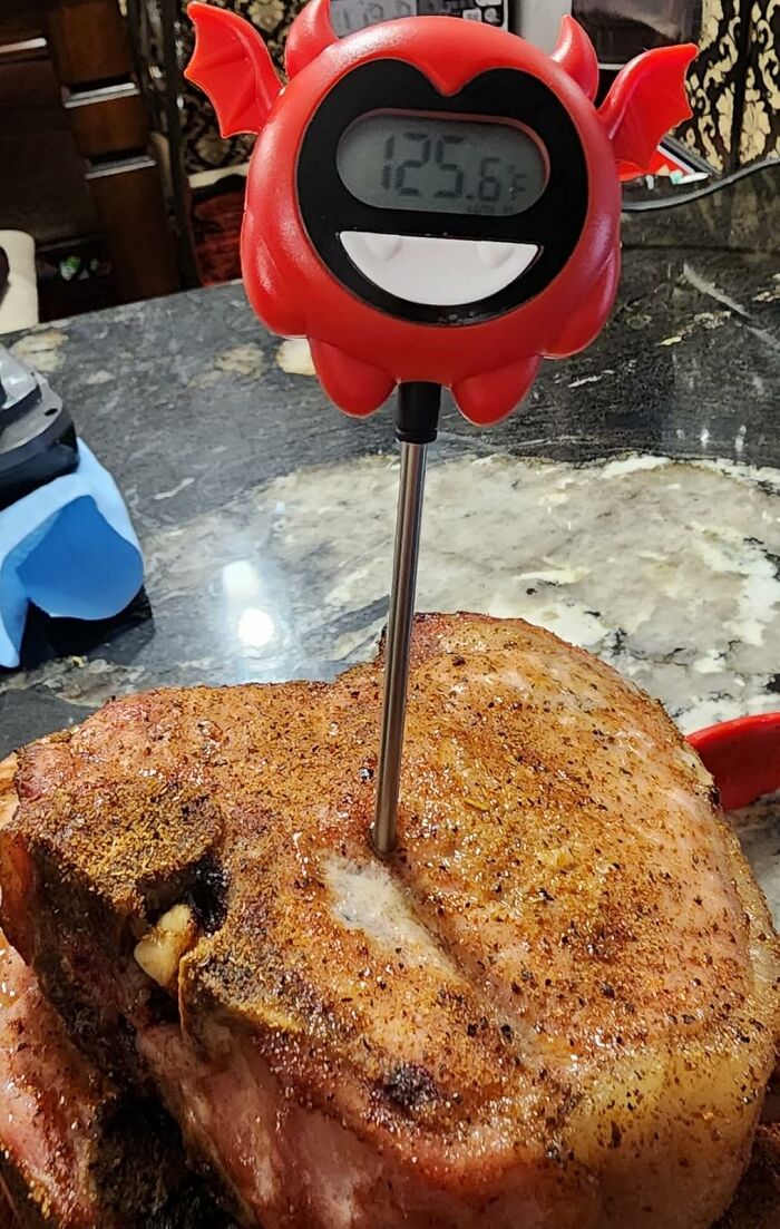 Red novelty meat thermometer with demon design inserted into seasoned cooked steak on a kitchen counter.
