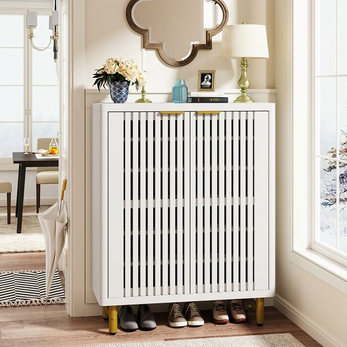 Modern white shoe cabinet with slatted doors and gold accents organizing a chaotic corner in a stylish home entryway.