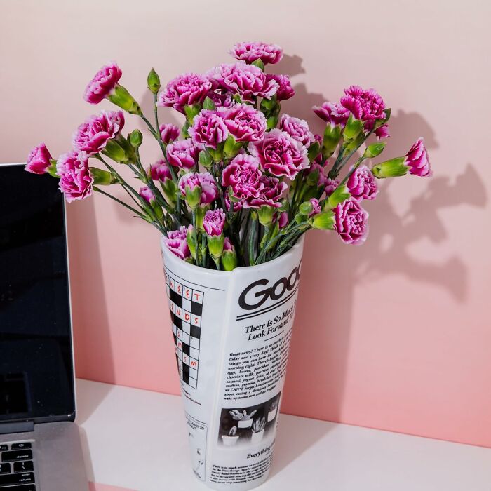 Pink carnations arranged in a retro newspaper-style vase beside a laptop on a pink background, vintage-inspired decor.