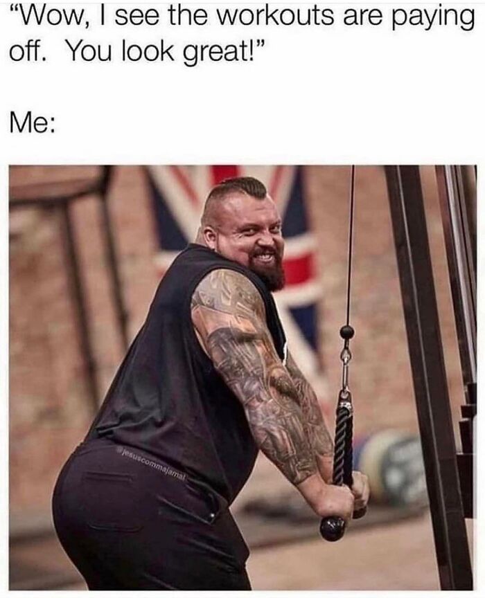 Muscular man with tattooed arms using gym cable machine, smiling humorously, representing gym life memes and workouts.