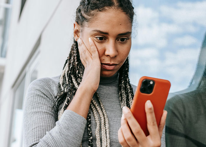 Woman rethinks her relationship with boyfriend looking worried and holding phone near a window in daytime setting