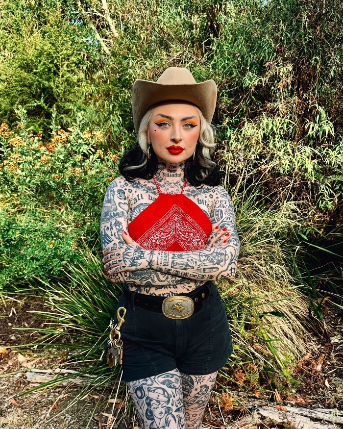Woman with full body suit tattoos wearing a red top and cowboy hat standing outdoors among greenery. - 4