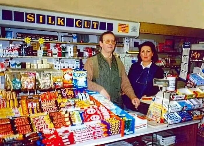 Two store clerks behind a counter filled with '90s candy and snacks, capturing nostalgic posts from growing up in the ’90s.