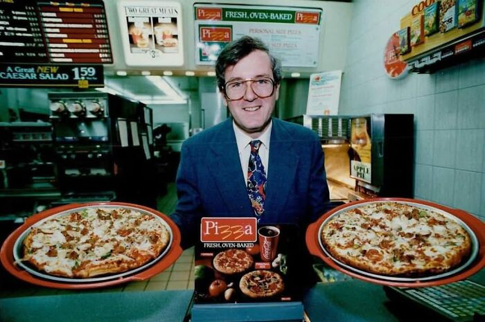 Man in a 90s pizza shop uniform holding two large fresh oven-baked pizzas, capturing 90s nostalgia and food culture.
