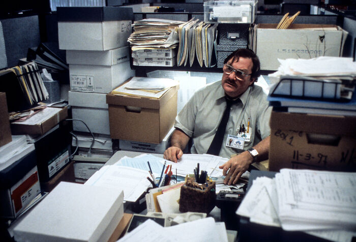 Man overwhelmed by cluttered office space filled with stacks of files and paperwork, illustrating consequences of job mistakes.