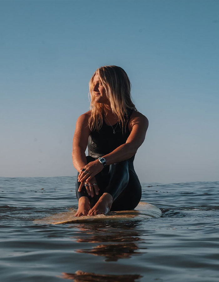 Surf instructor sitting on a surfboard in the ocean during sunset, wearing a black wetsuit and looking to the side.